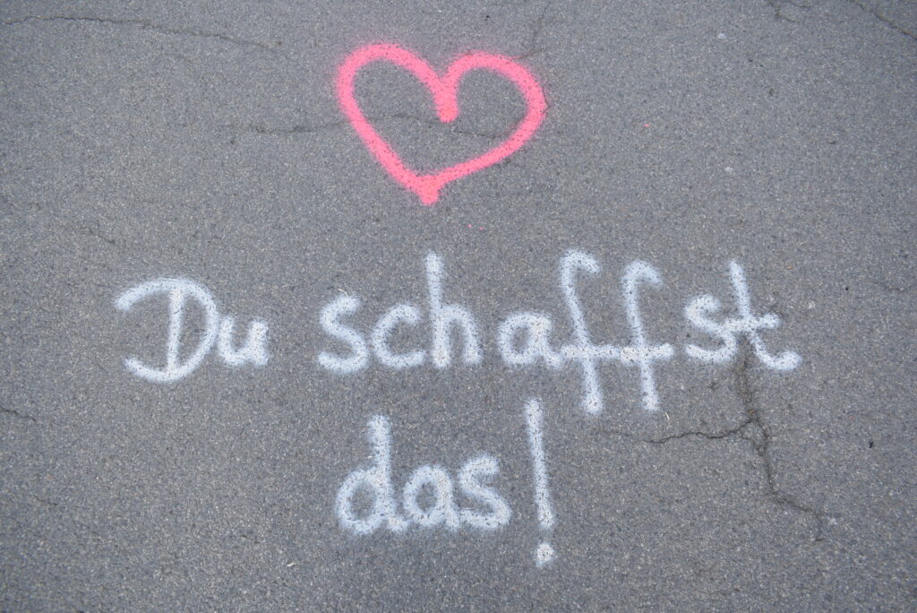Mit weißer Sprühkreide steht auf Asphalt geschrieben: "Du schaffst das". Über den Schriftzug ist noch ein pink-farbiges Herz gezeichnet.