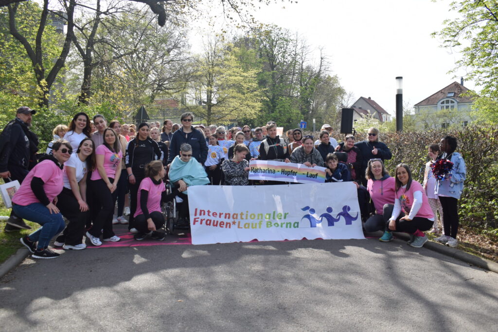 An einer Startlinie aufgreiht stehen und sitzen in Rollis aufgreiht verschiedene Frauen und halten ein Banner mit der Aufschrift "Internationaler Frauen*Lauf Borna" sowie "Tirhas-Fishaye-Lauf" in die Kamera. Die Stimmung scheint freudig und ausgelassen.