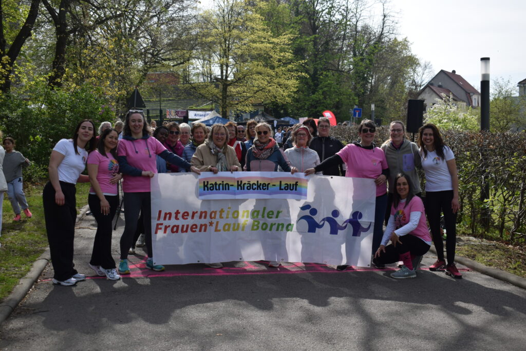 An einer Startlinie stehen aufgereiht verschiedene Frauen und Kinder und halten ein Banner mit der Aufschrift "Internationaler Frauen*Lauf Borna" sowie "Katrin-Kräcker-Lauf" in die Kamera. Die Stimmung scheint freudig und ausgelassen.