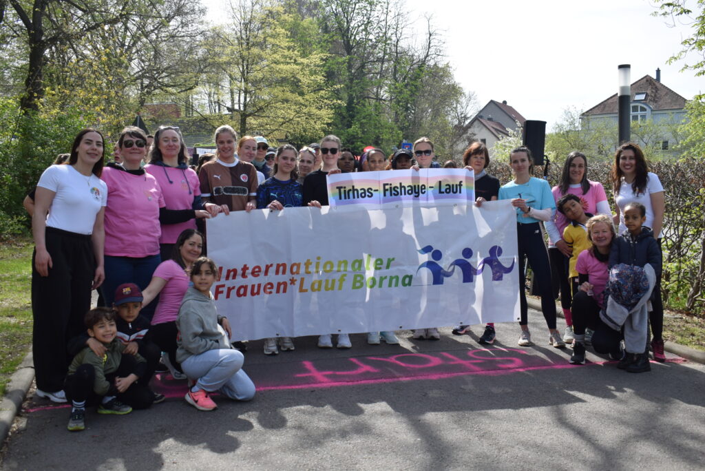 An einer Startlinie aufgreiht stehen aufgreiht verschiedene Frauen und Kinder und halten ein Banner mit der Aufschrift "Internationaler Frauen*Lauf Borna" sowie "Tirhas-Fishaye-Lauf" in die Kamera. Die Stimmung scheint freudig und ausgelassen.