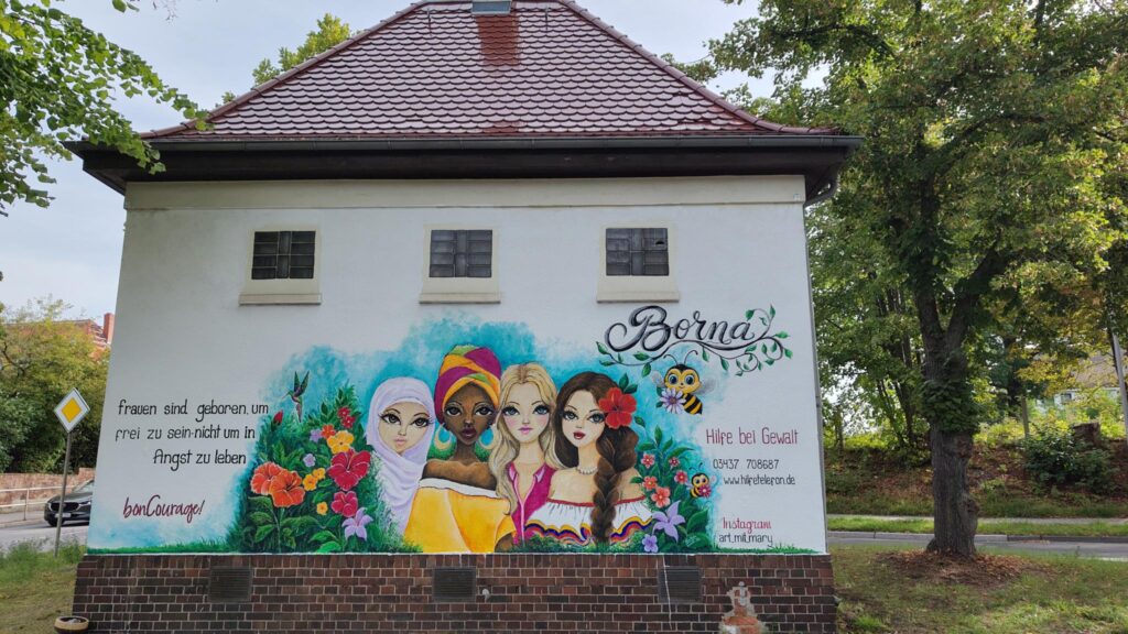An der Wand eines kleines Häuschen ist ein sehr farbenfrohes Bild gemalt, in dessen Zentrum vier verschiedene Frauen sind, umrahmt von Grün und Blumen. Zudem finden sich verschiedene Schriftzüge, u.a. "Frauen sind geboren, um frei zu sein - nicht um in Angst zu leben" oder "wee.hilfetelefon.de".